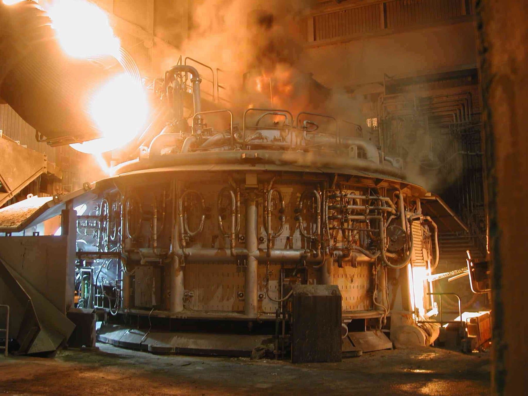 An electric arc furnace in operation, the charge glowing inside the shop.